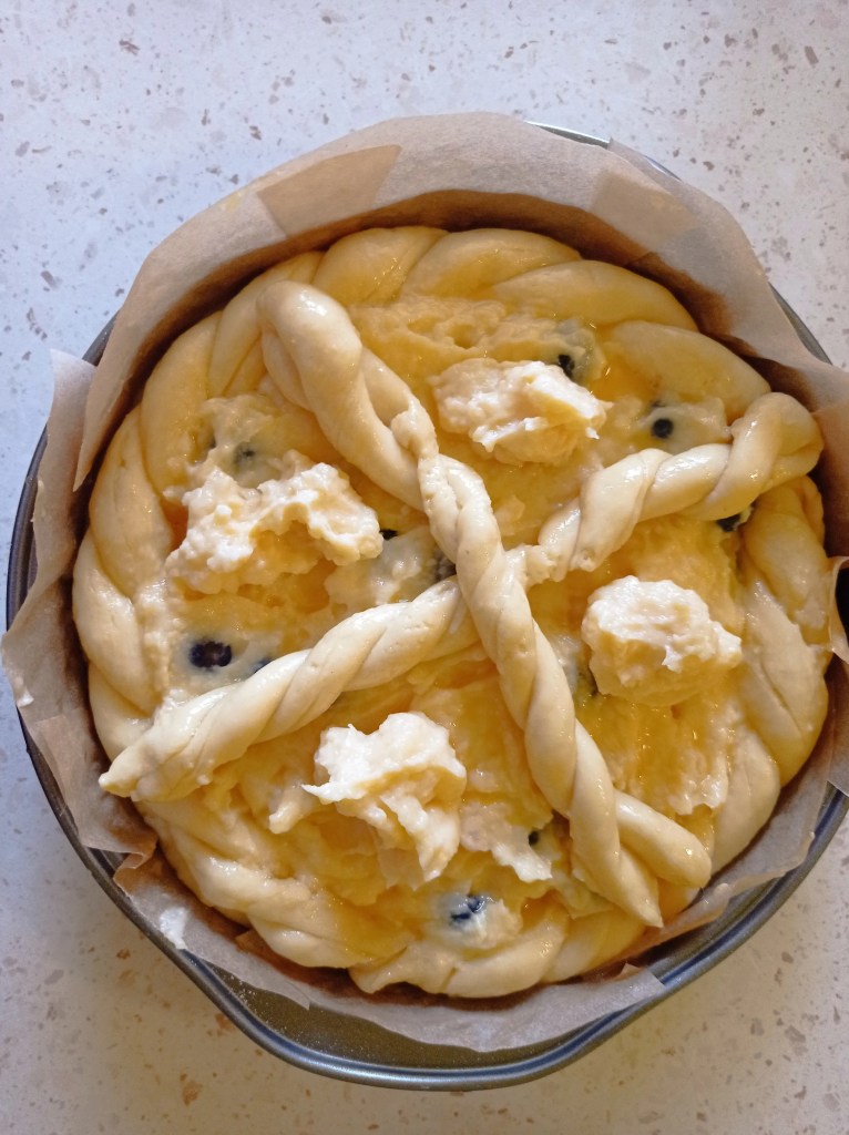 Unbaked dairy-free Pasca with visible vanilla cream dollops and fresh blueberries, decorated with a twisted dough cross