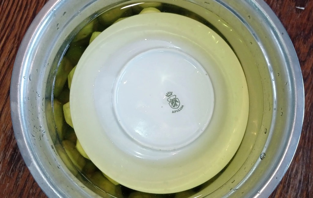 Peeled green walnuts submerged in water, weighted down with a plate before refrigeration.