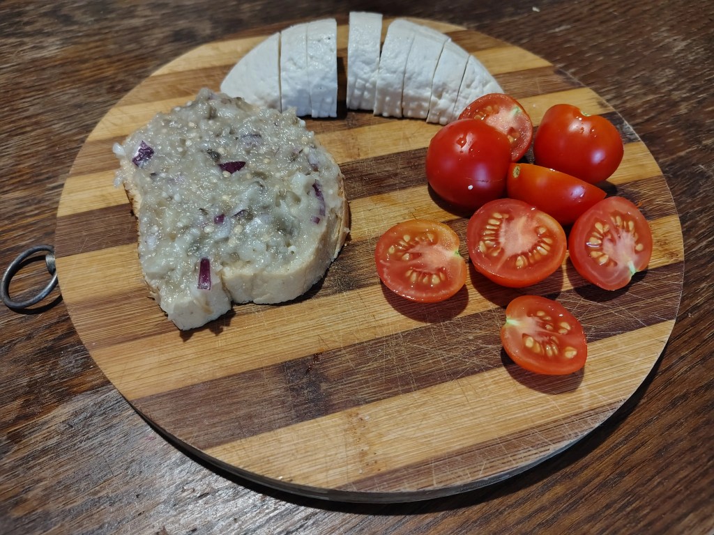 slice of bread topped with Romanian eggplant salad, served alongside sliced cheese and fresh cherry tomatoes on a wooden board