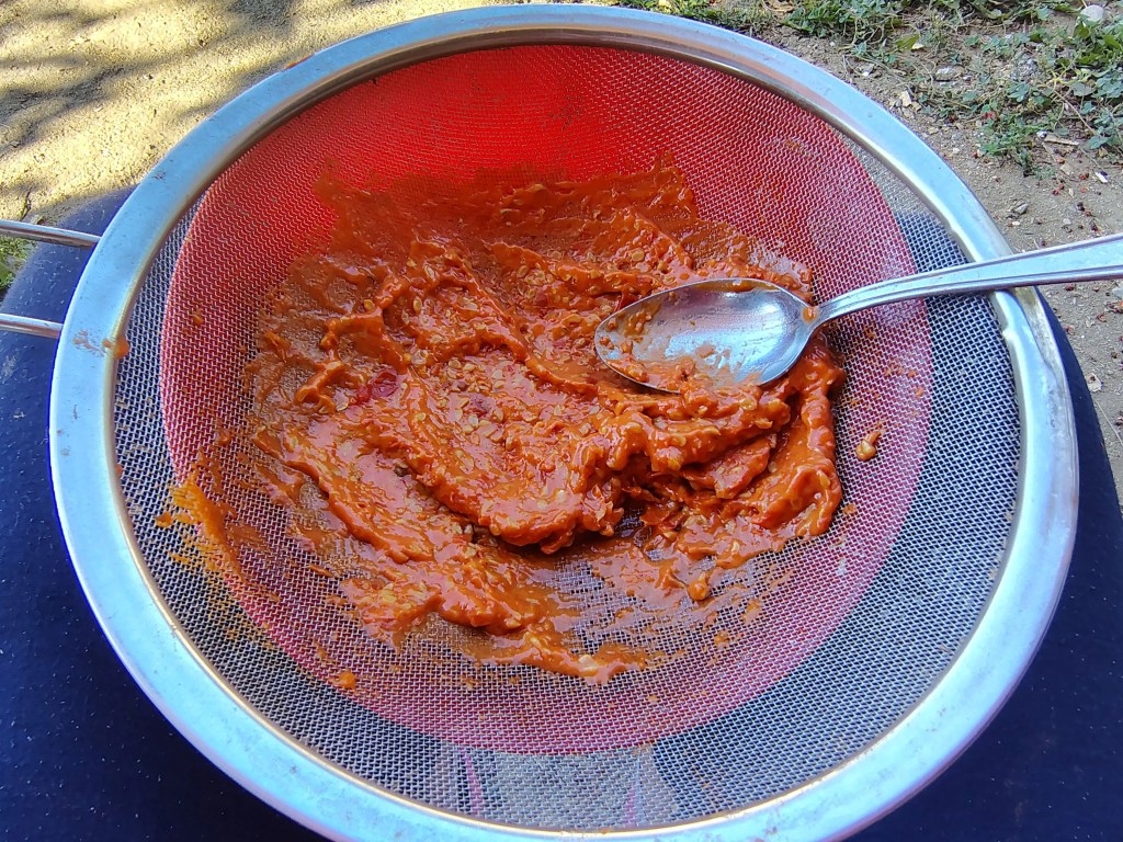 Rosehip pulp being pressed through a fine sieve using a spoon during the jam-making process.